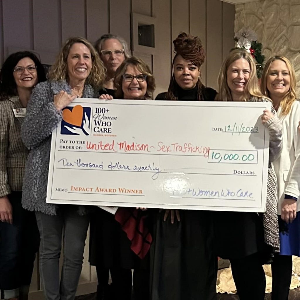 A group of 100+ Women Who Care - Madison members gather around a charity presenter receiving the Impact Award oversize check.
